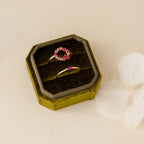 Two gold rings with pink stones sit in a 2 Slot Velvet Ring Box made of elegant green velvet, displayed beside a white flower on a cream surface.