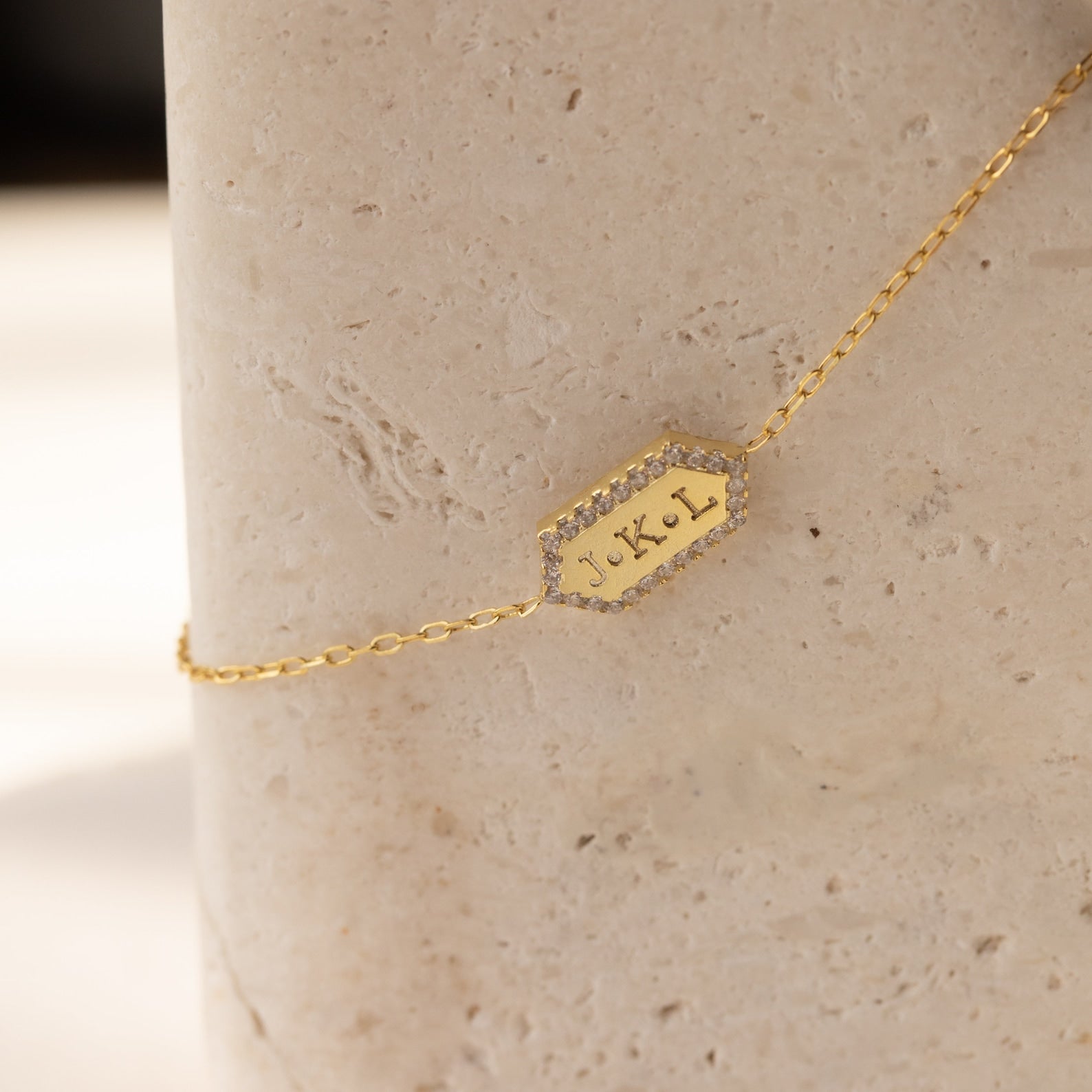 The Custom Pave Hexagon Bracelet features a gold chain and a hexagonal charm engraved with J + L, accented with small crystals—ideal for anniversaries. Shown on a stone background.