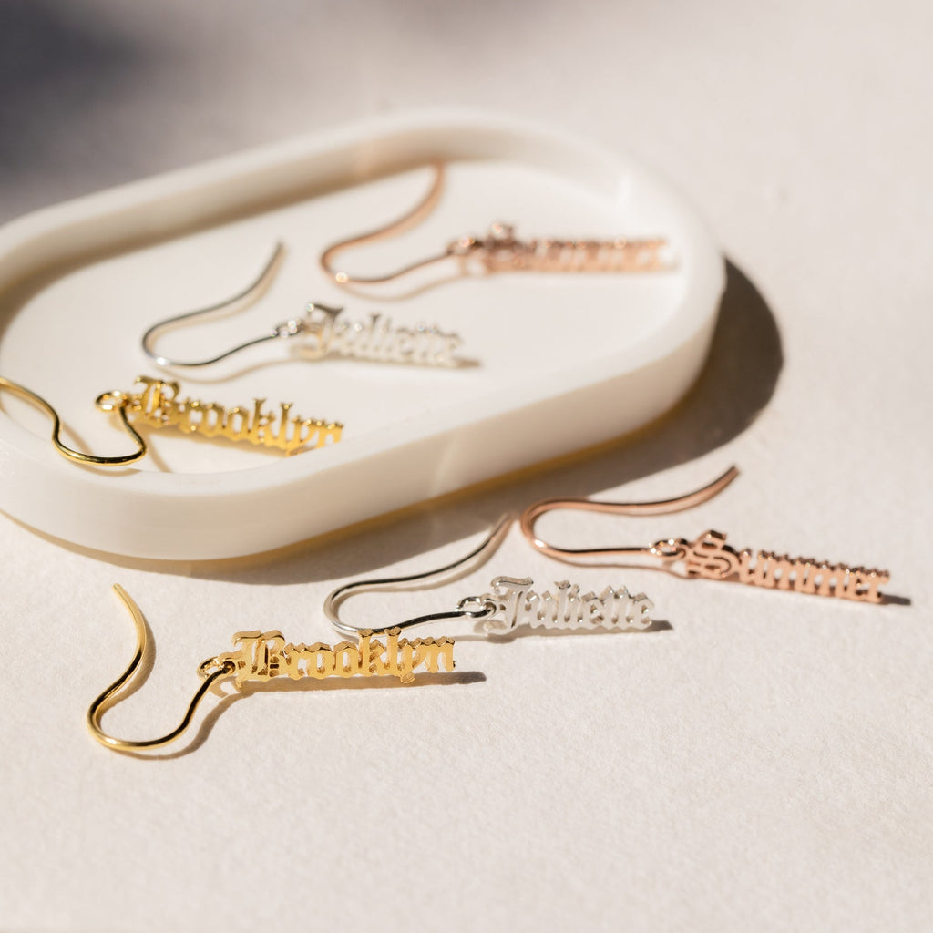 Old English Dangling Name Earrings in gold and silver with different names are shown on a white oval dish, making them a stylish handmade drop earring and perfect custom gift.