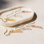 Old English Dangling Name Earrings in gold and silver with different names are shown on a white oval dish, making them a stylish handmade drop earring and perfect custom gift.