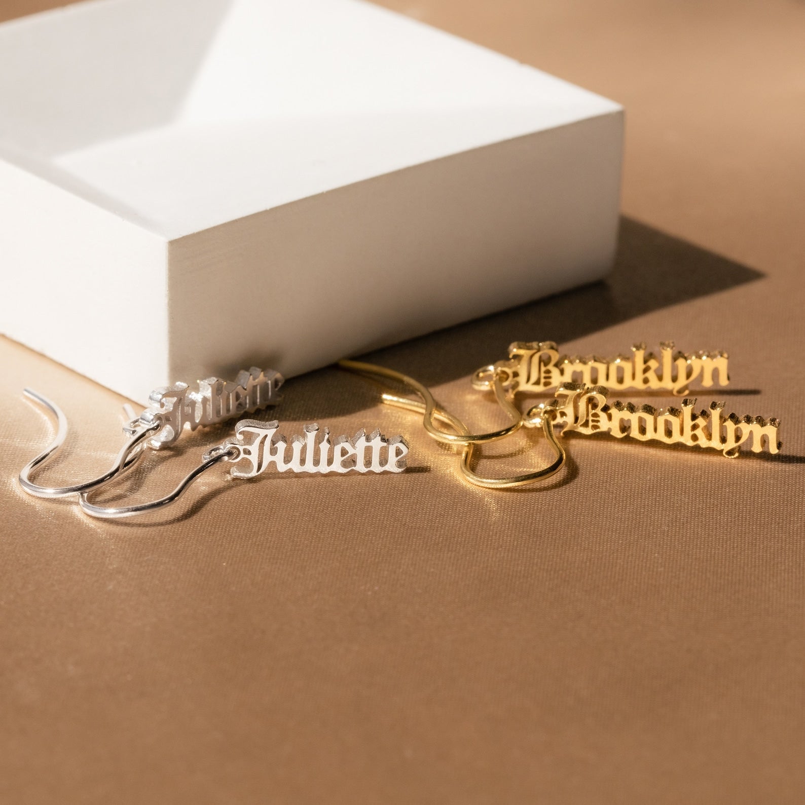 Four Old English Dangling Name Earrings in silver and gold, customized with “Juliette” and “Brooklyn,” displayed on a tan surface—ideal as a personalized gift or for those wanting handmade drop earrings.