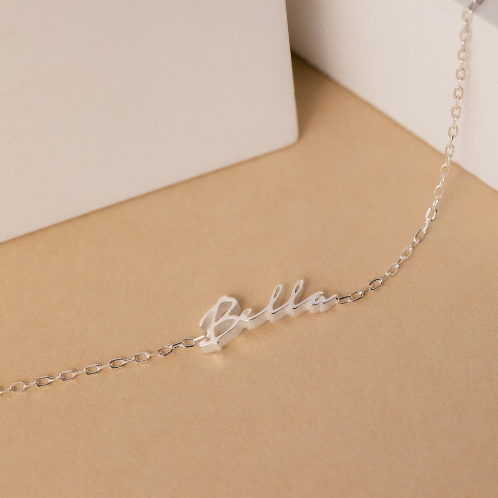 Displayed on a beige background, this silver chain necklace features a cursive "Bella" pendant—a perfect delicate accent for minimal jewelry lovers or anyone looking to pair it with their Venice Tiny Name Bracelet.