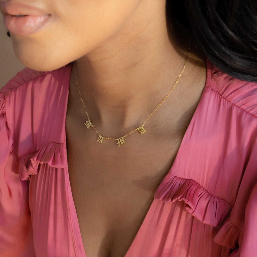 A woman wears a pink blouse and an Old English Letter Name Necklace with small letter charms around her neck.