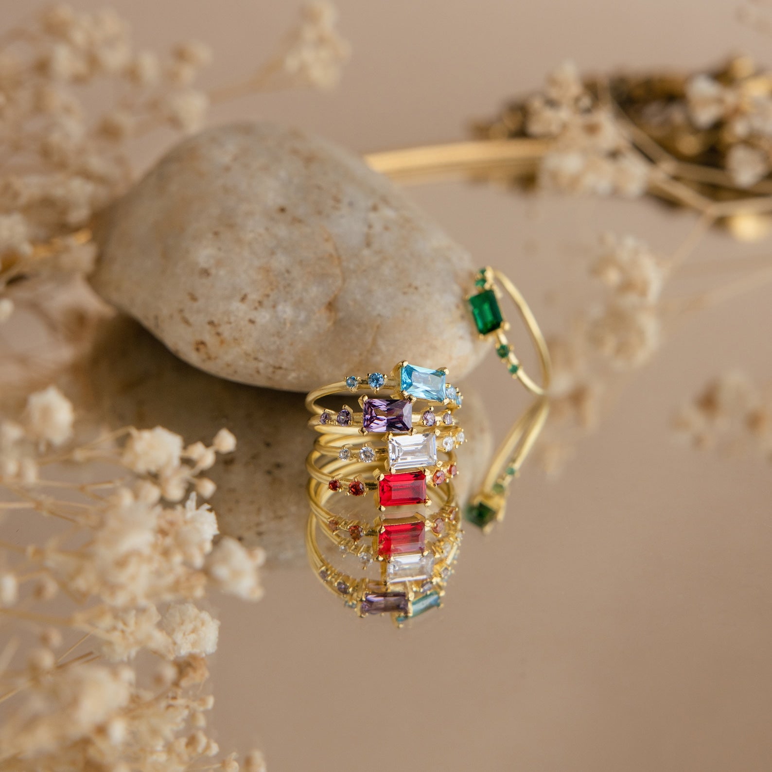 The Space Baguette Birthstone Rings are stacked on a stone, reflected on a mirrored surface with dried flowers.