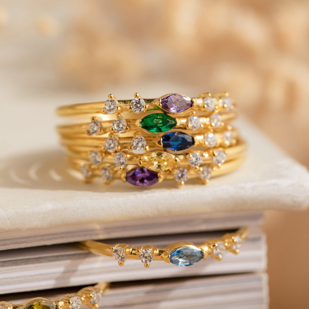 Stack of gold rings with crystal stones, colorful gemstones, and small diamonds—featuring the customizable Space Marquise Birthstone Ring—artfully displayed on a white book.