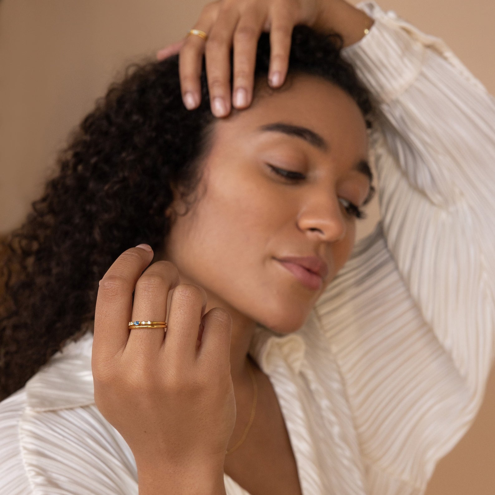 A woman in a white blouse poses with her hand near her face, highlighting gold rings including the Space Marquise Birthstone Ring adorned with sparkling crystal stones.