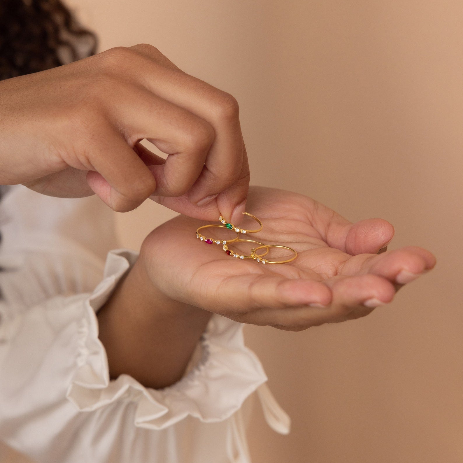 A person holds several Space Marquise Birthstone Rings, featuring delicate gold bands with colorful crystal stones and customizable gemstone accents, against a neutral background.