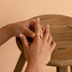 A person slips a Bold Birthstone Signet Ring in Rose Gold onto their finger beside a wooden stool.