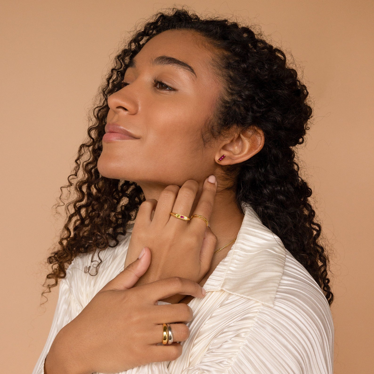 A woman with curly hair wears the Bold Birthstone Signet Ring in Rose Gold and gold earrings, touching her neck and gazing to the side against a beige background.