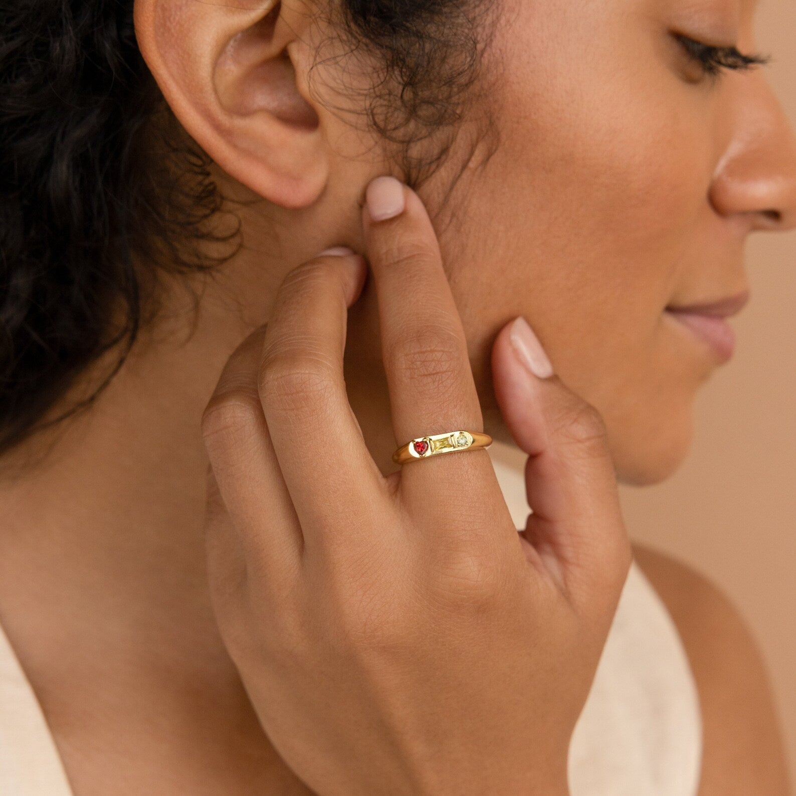 A woman touches her face, wearing the Bold Birthstone Signet Ring adorned with colorful gemstones on her finger.
