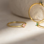 The Tiny Heart Birthstone Ring is a delicate gold heart-shaped ring with a small pink gemstone, resting on a light surface—a perfect romantic gift.