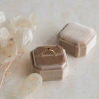 The Tiny Heart Birthstone Ring features a gold heart design with a small red stone, presented in a beige velvet box beside pale dried leaves—a delicate and romantic gift.