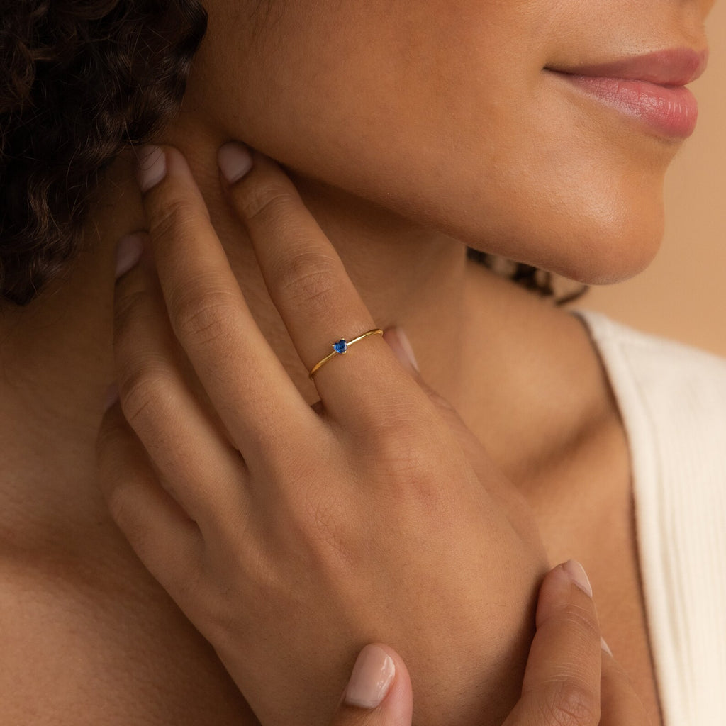 A woman gently touches her neck, wearing the Tiny Heart Birthstone Ring—a delicate gold stacking ring with a small blue stone—on her finger.