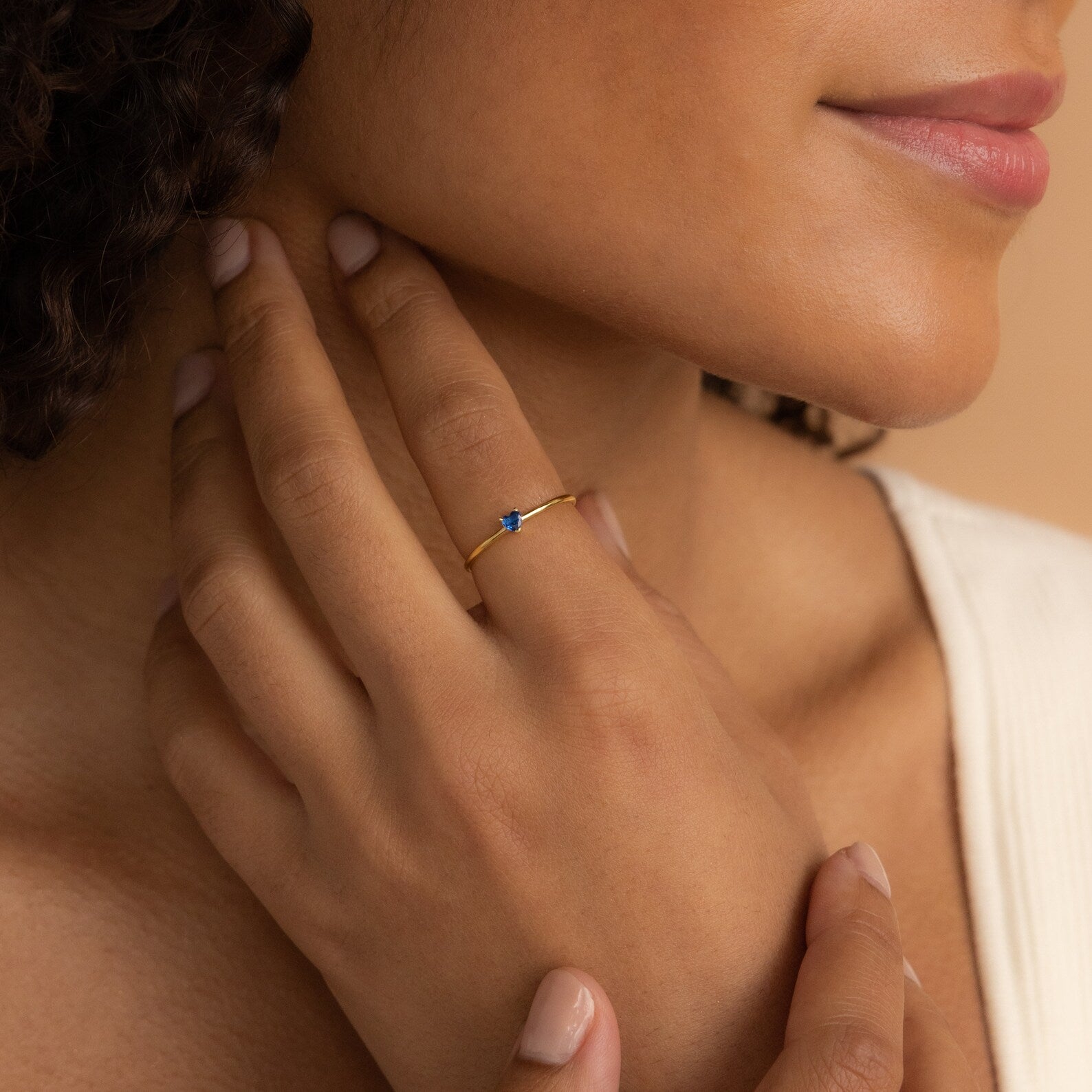 A woman gently touches her neck, wearing the Tiny Heart Birthstone Ring—a delicate gold stacking ring with a small blue stone—on her finger.