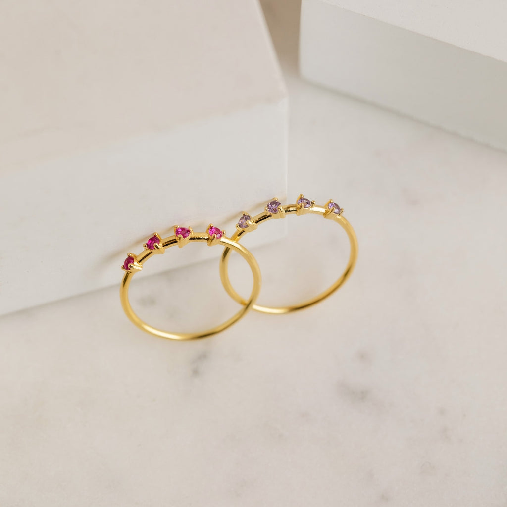 Two Space Birthstone Rings with small pink and clear stones are displayed on a white marble surface near white blocks—a perfect stackable multi-stone ring set.