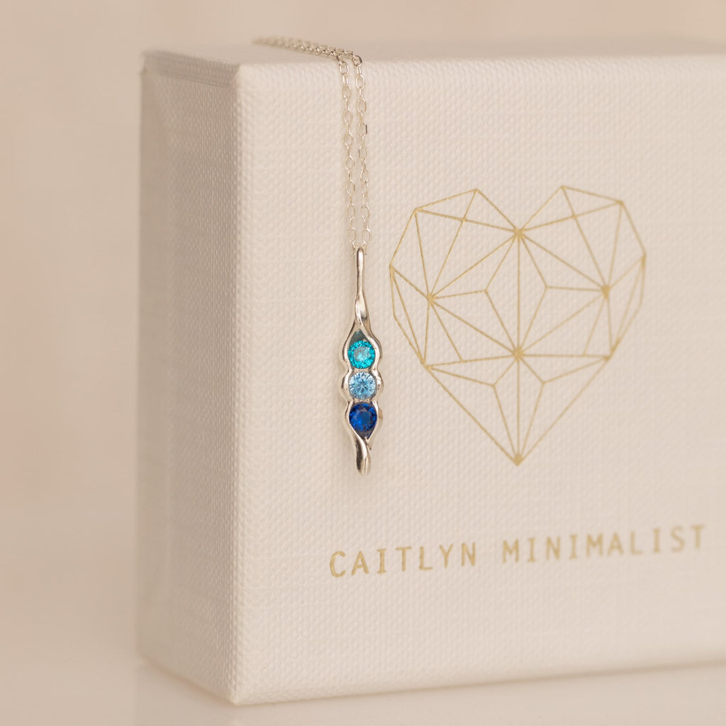 The Vertical Pod Birthstone Necklace features a silver pendant with three blue gemstones on a chain, displayed against a white box with a gold geometric heart—perfect for personalized jewelry lovers.