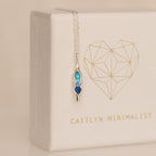 The Vertical Pod Birthstone Necklace features a silver pendant with three blue gemstones on a chain, displayed against a white box with a gold geometric heart—perfect for personalized jewelry lovers.