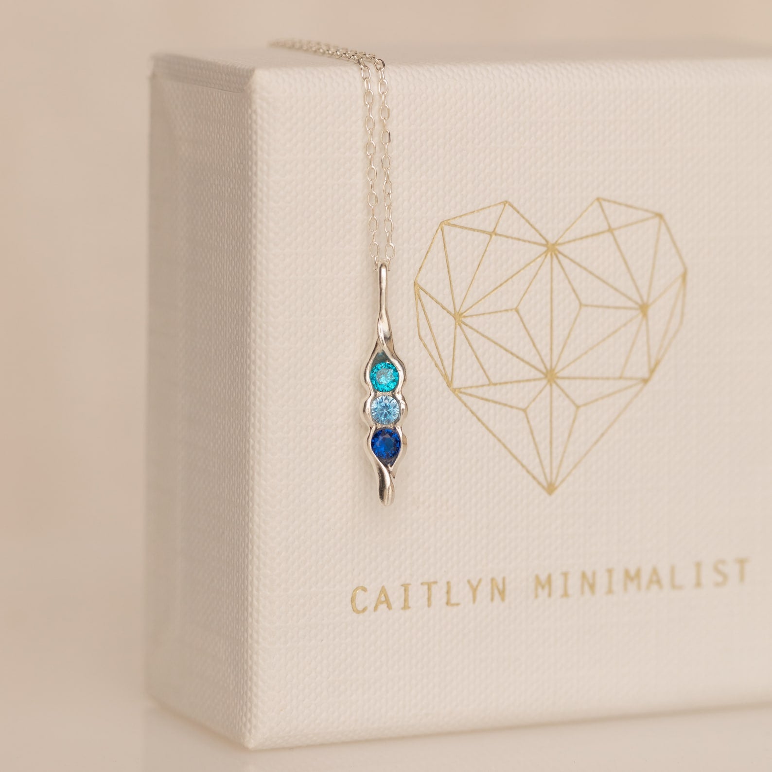 The Vertical Pod Birthstone Necklace features a silver pendant with three blue gemstones on a chain, displayed against a white box with a gold geometric heart—perfect for personalized jewelry lovers.