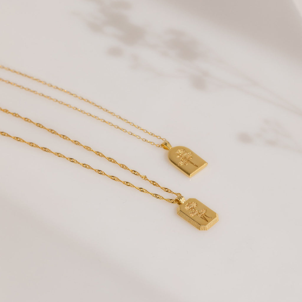 On a light surface with soft floral shadows, the Ribbed Flower Tag Necklace exudes timeless charm and subtle elegance, evoking vintage-inspired style.