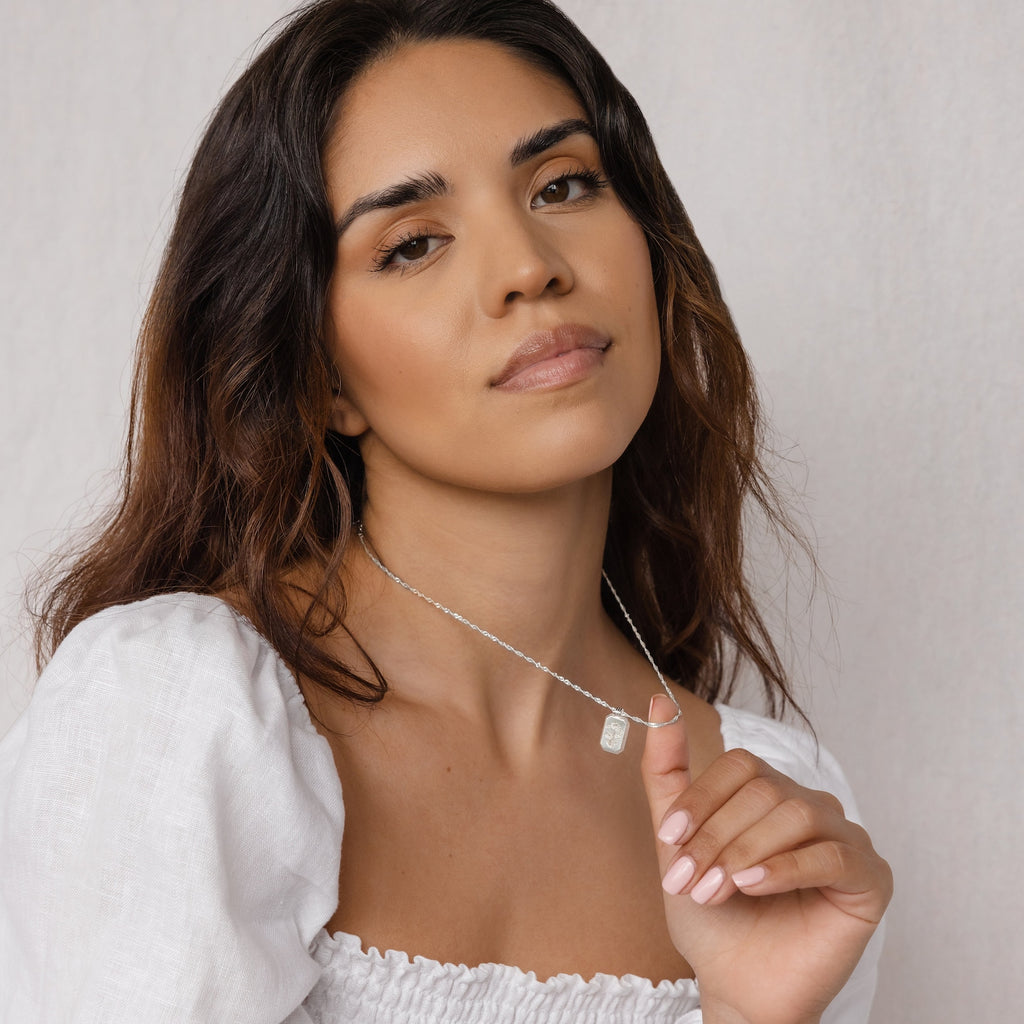 A woman in a white top looks at the camera while holding the Ribbed Flower Tag Necklace against a light background.