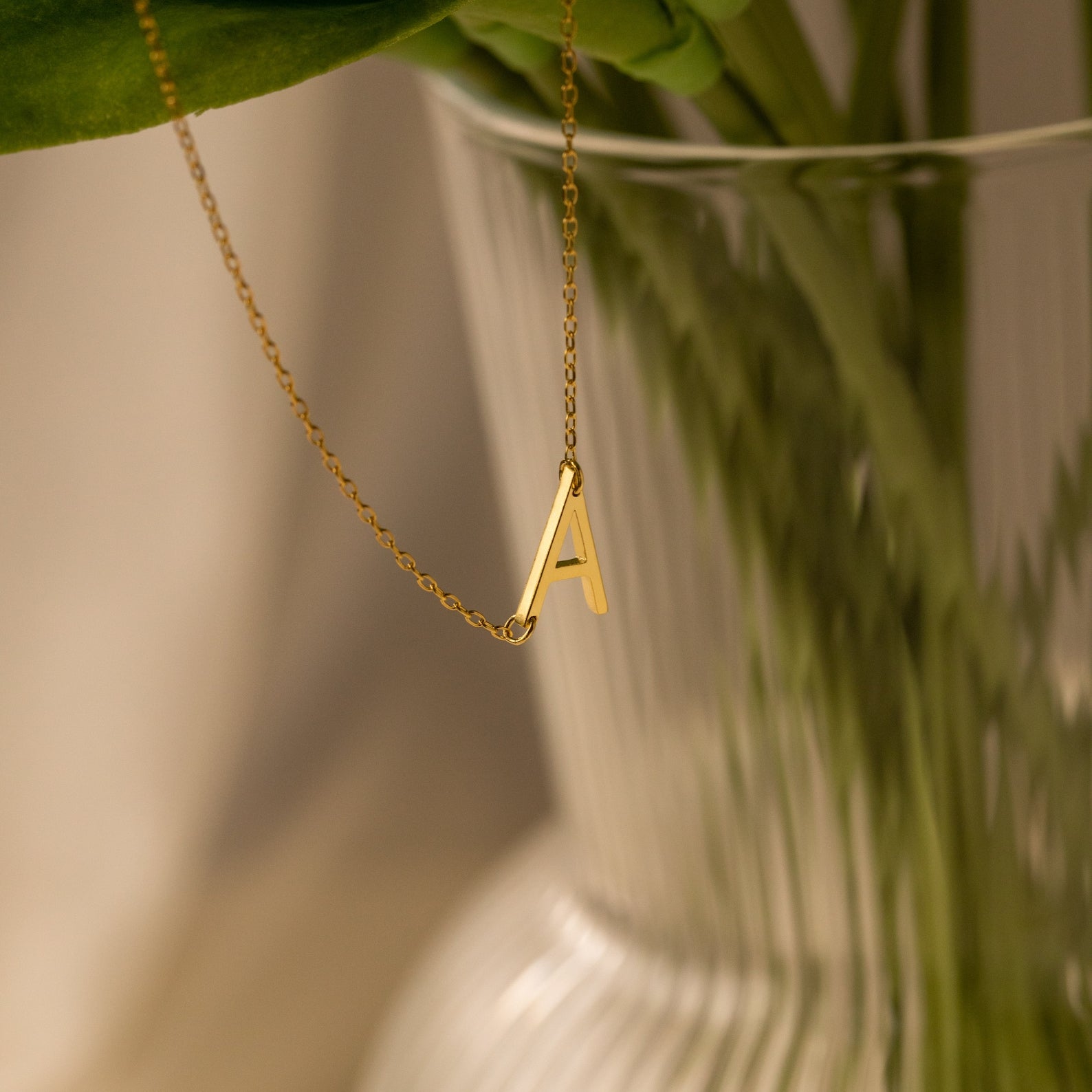 The Mini Sideways Initial Necklace, featuring a gold letter A pendant, drapes elegantly over a clear ribbed glass vase—making it an ideal personalized gift for any occasion.