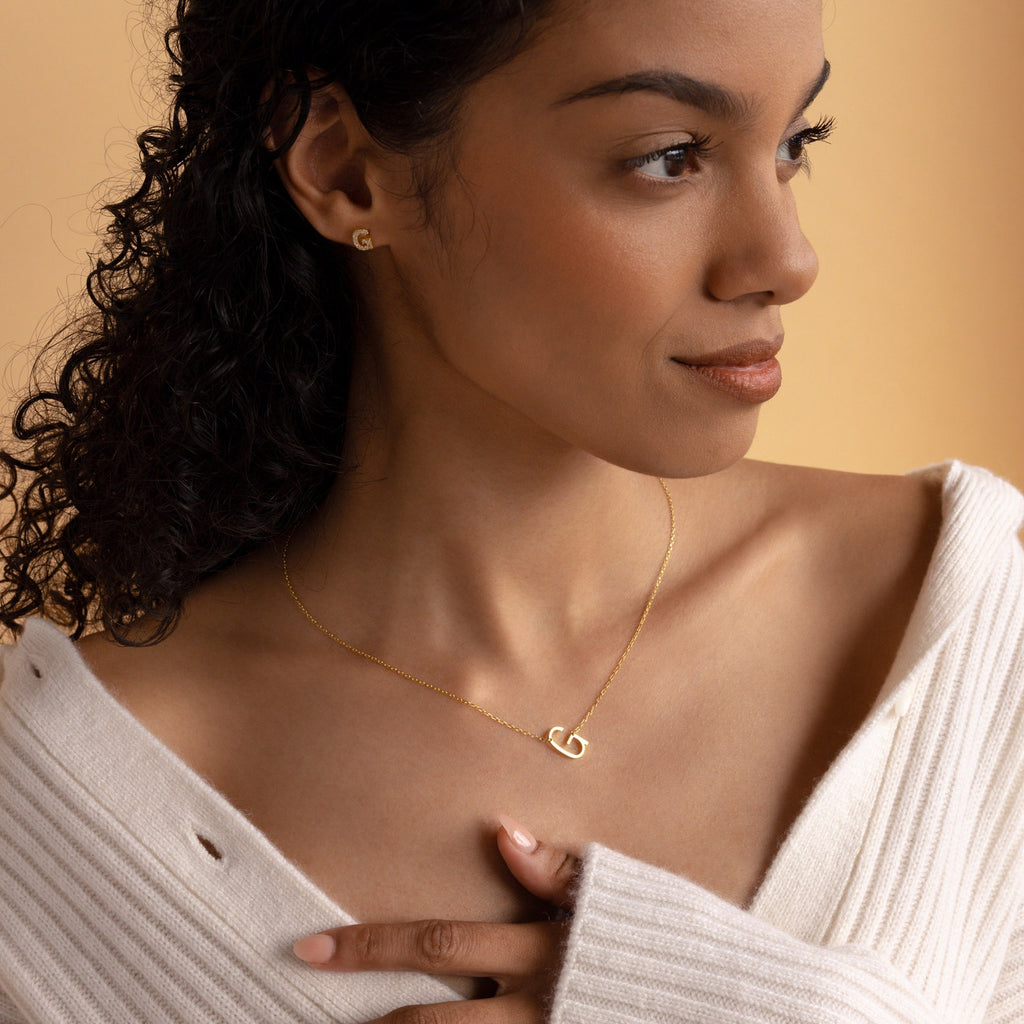 A woman in a cream sweater looks to the side, wearing the Mini Sideways Initial Necklace and earrings—a perfect personalized gift.