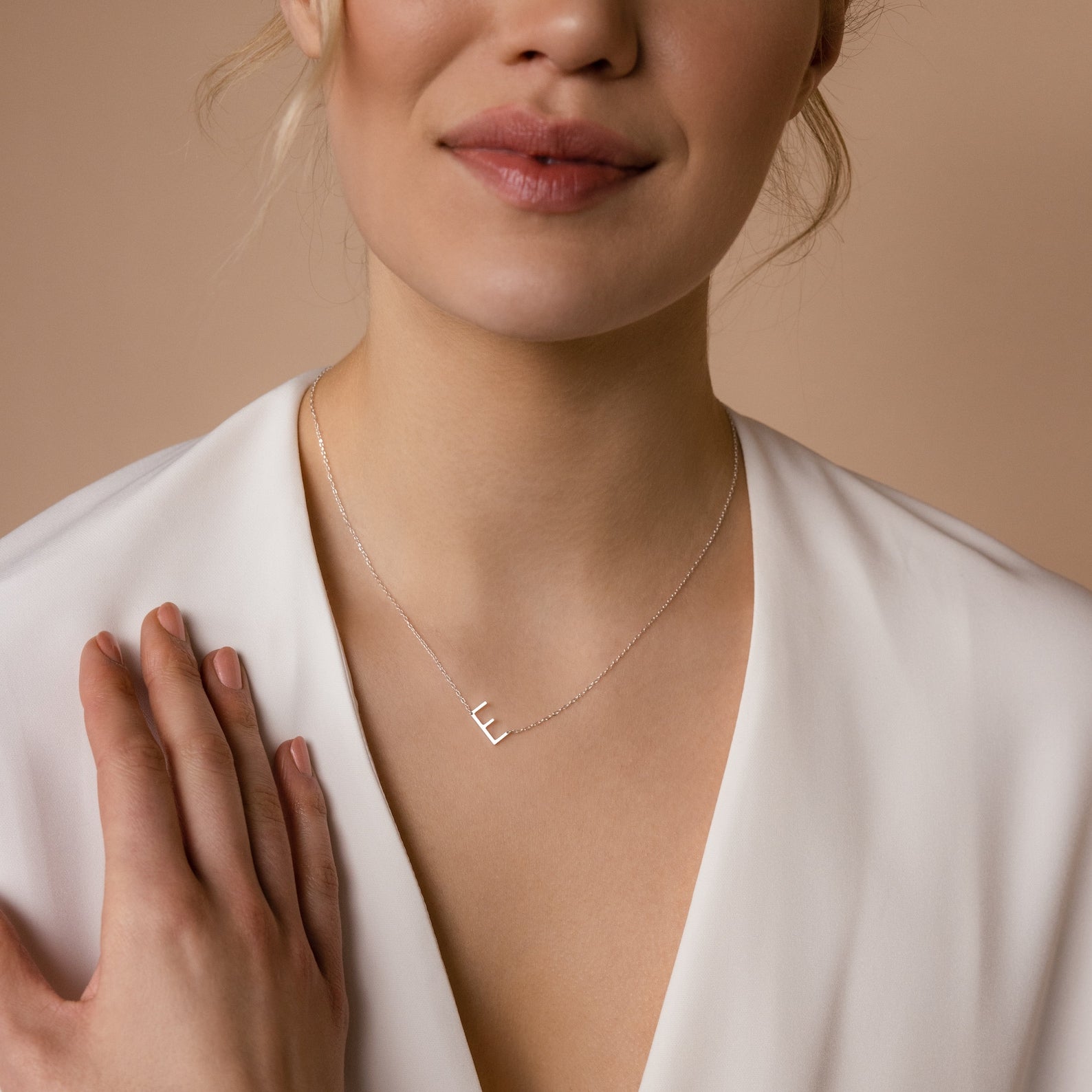 A woman in a white blouse wears the Mini Sideways Initial Necklace, featuring a delicate silver horizontal “L” pendant—a chic and thoughtful personalized gift.