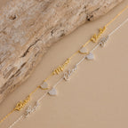 A gold and a silver nameplate bracelet with heart charms and cursive names rest on beige, ideal for pairing with your Grace Name Heart Necklace.