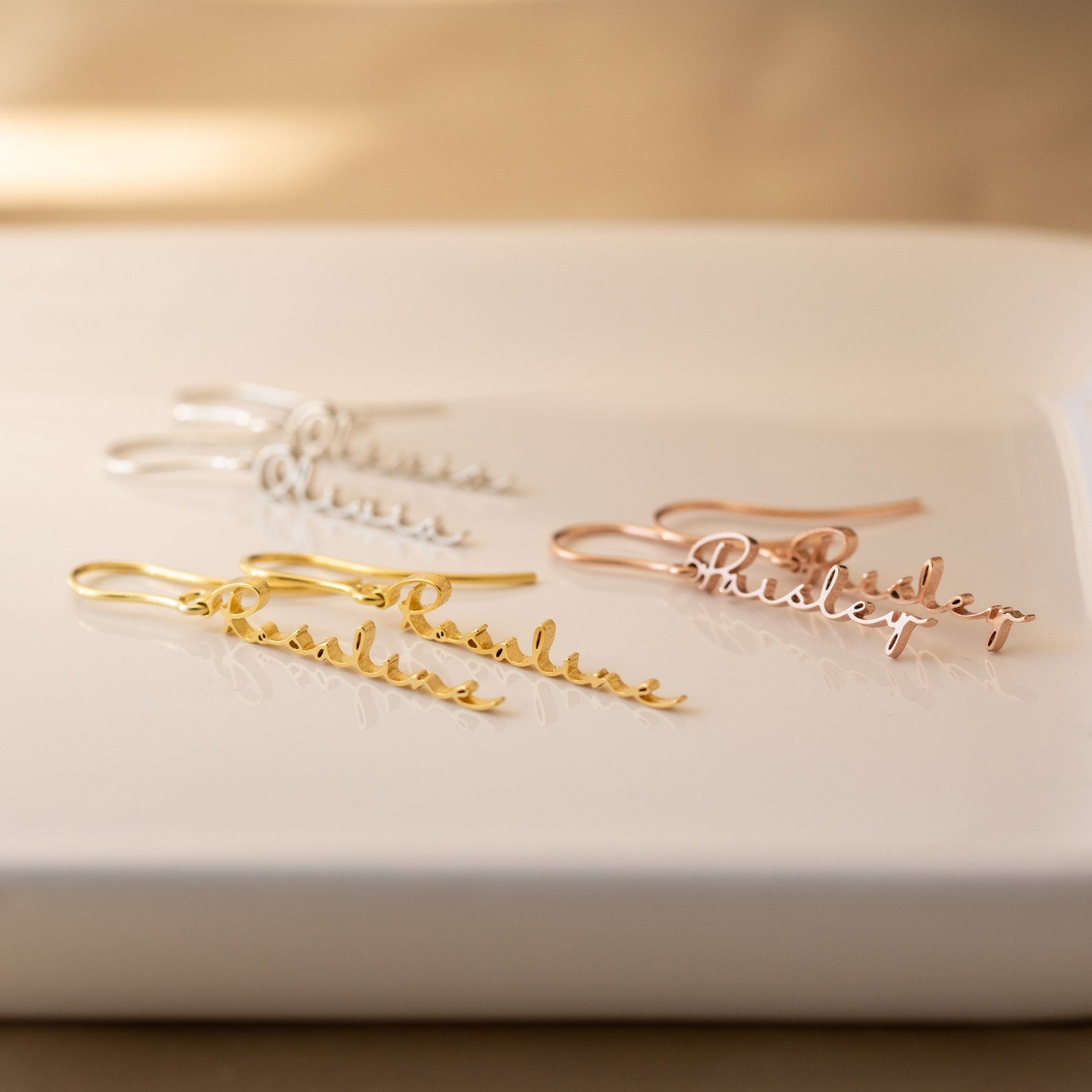 Add a special touch to any look with three pairs of Hallie Dangling Name Earrings in gold, silver, and rose gold—perfect for fans of personalized jewelry. Shown beautifully displayed on a white surface.