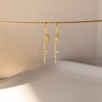 Hallie Dangling Name Earrings hang delicately on a thin wire against a softly lit neutral background, highlighting the elegance of personalized name jewelry.
