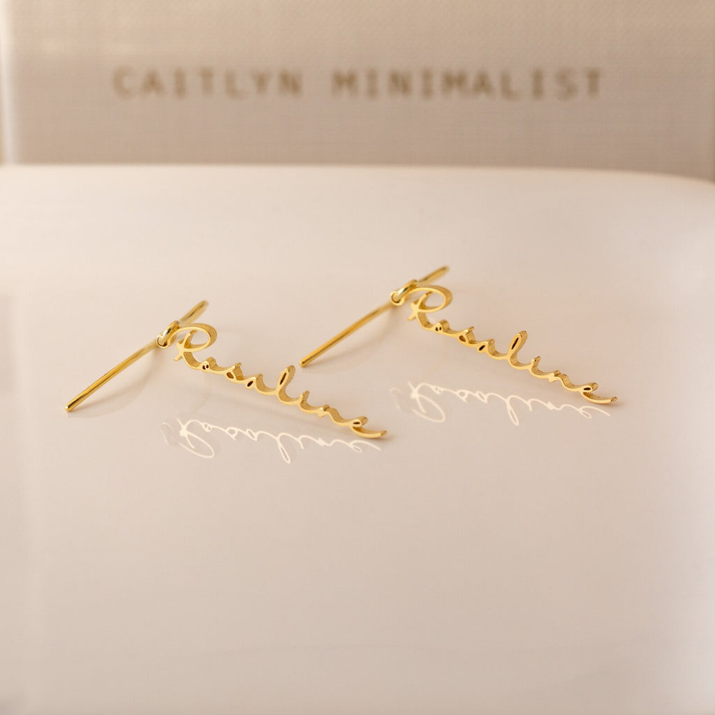 Hallie Dangling Name Earrings spelling Pauline in cursive rest on a white surface, with CAITLYN MINIMALIST in the background—a chic piece of personalized jewelry perfect for any style.