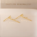 Hallie Dangling Name Earrings spelling Pauline in cursive rest on a white surface, with CAITLYN MINIMALIST in the background—a chic piece of personalized jewelry perfect for any style.