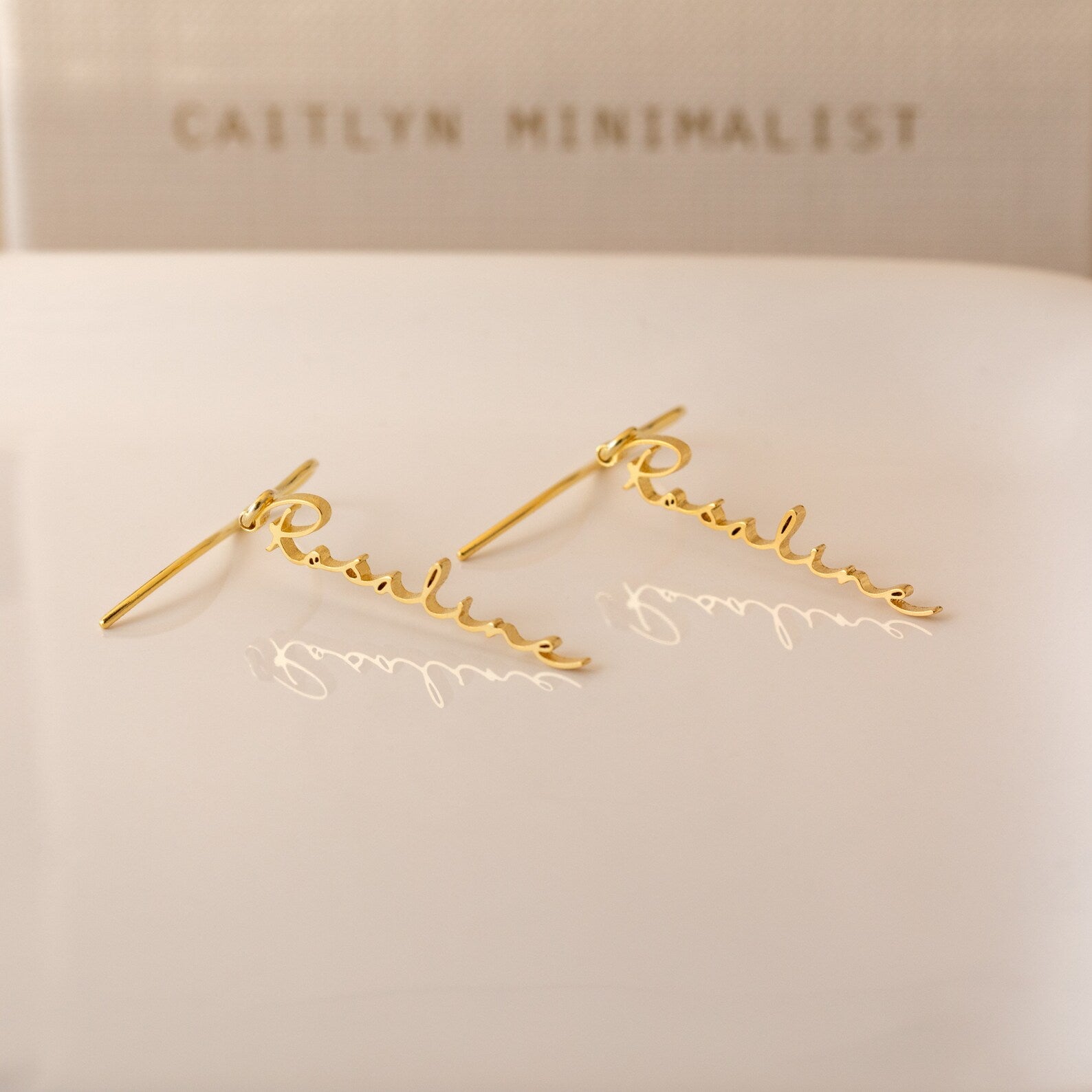 Hallie Dangling Name Earrings spelling Pauline in cursive rest on a white surface, with CAITLYN MINIMALIST in the background—a chic piece of personalized jewelry perfect for any style.