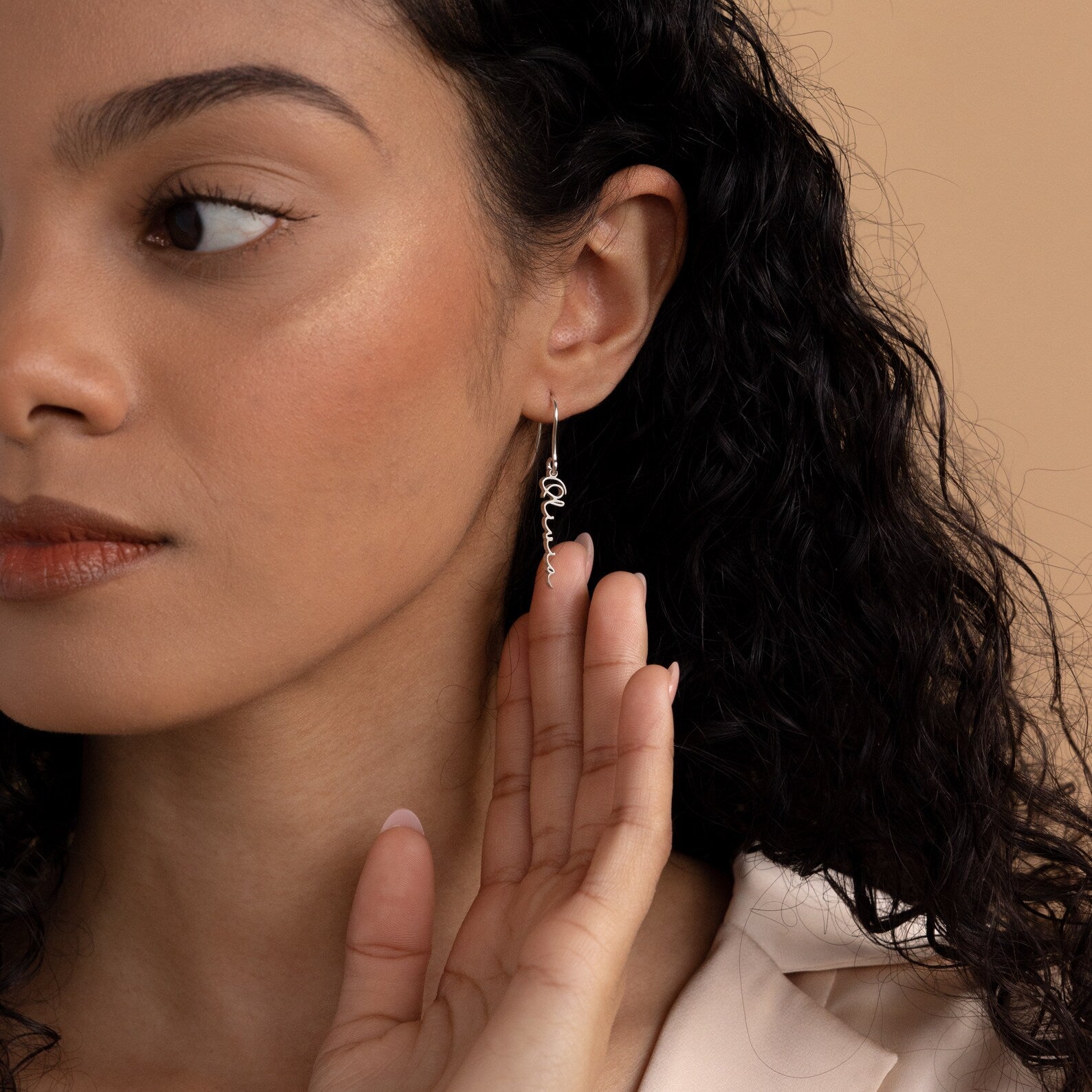 A woman with curly hair touches her Hallie Dangling Name Earring while looking to the side against a neutral background.