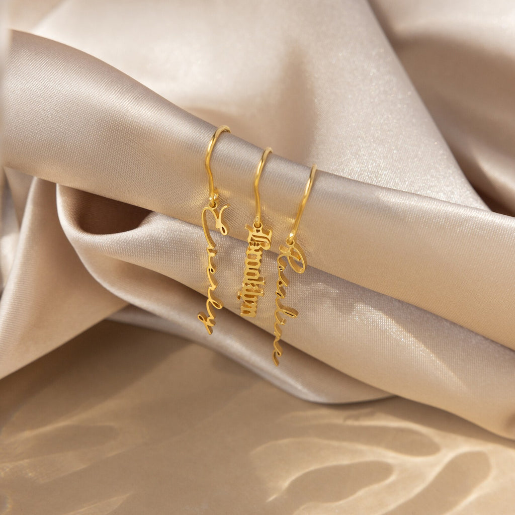 Three Hallie Dangling Name Earrings with gold hoops and personalized name charms, a striking example of custom jewelry, displayed on soft, champagne-colored satin fabric.