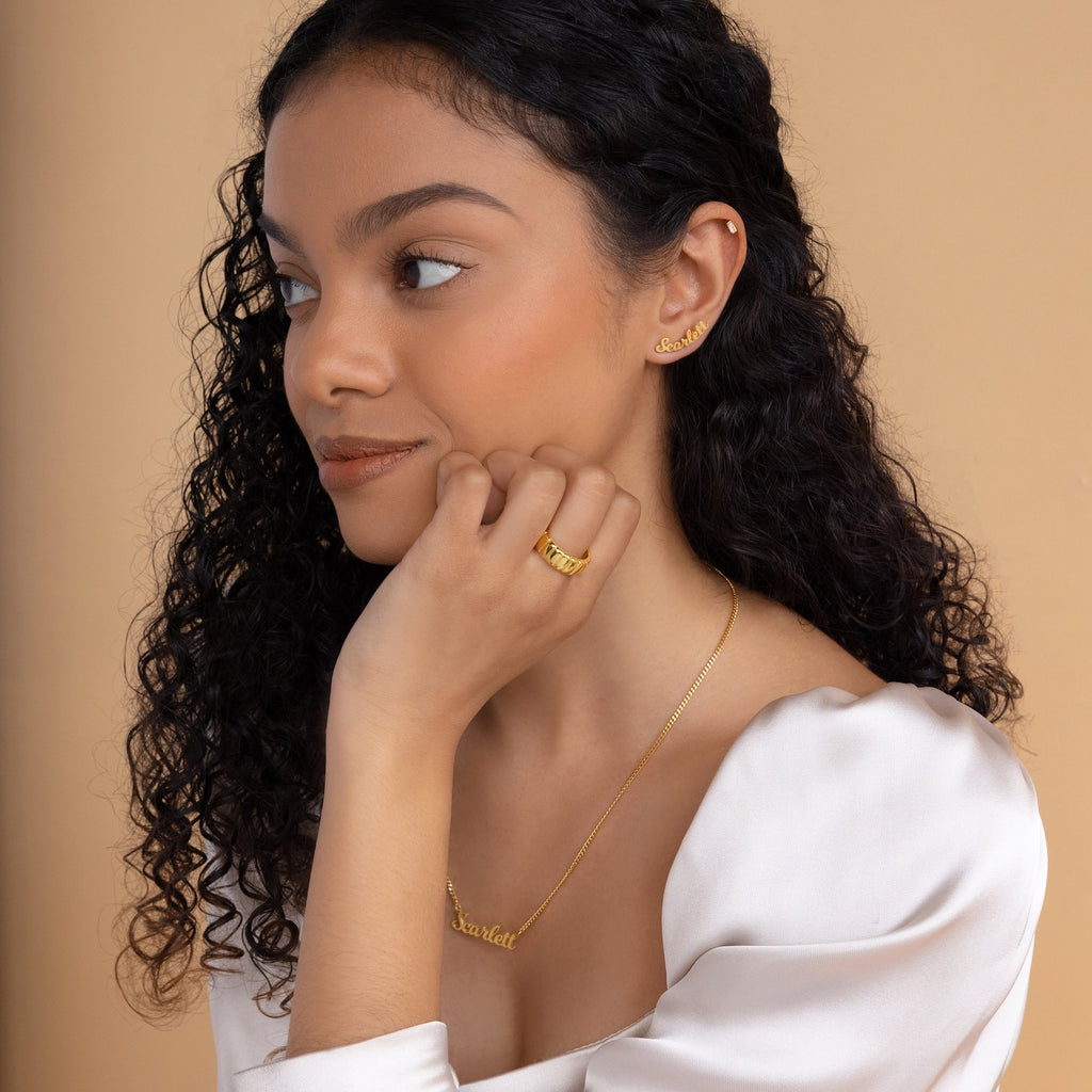 Woman with curly hair wears Mellow Name Earrings and gold name jewelry, paired with a white top, looking to the side against a beige background.
