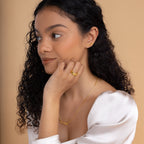 Woman with curly hair wears Mellow Name Earrings and gold name jewelry, paired with a white top, looking to the side against a beige background.