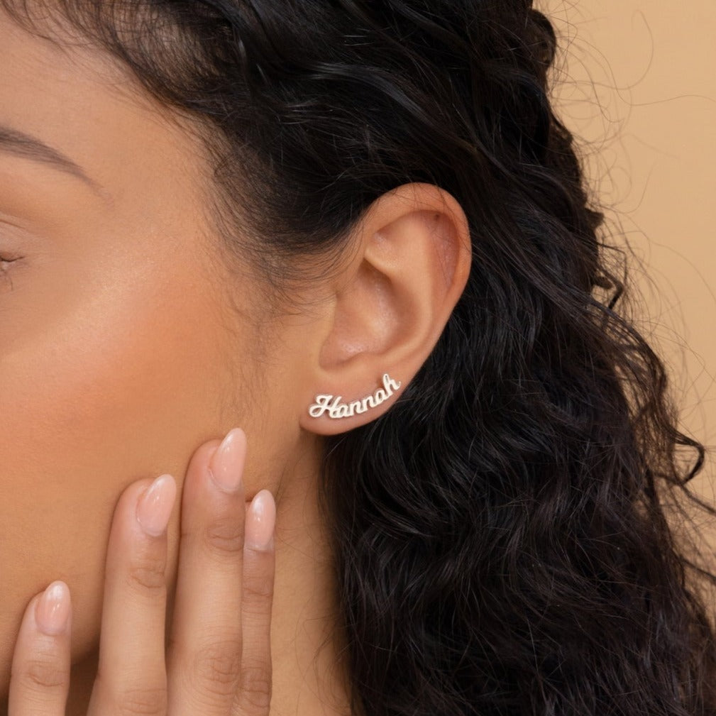 A woman with curly hair wears Mellow Name Earrings and touches her face with manicured fingers.