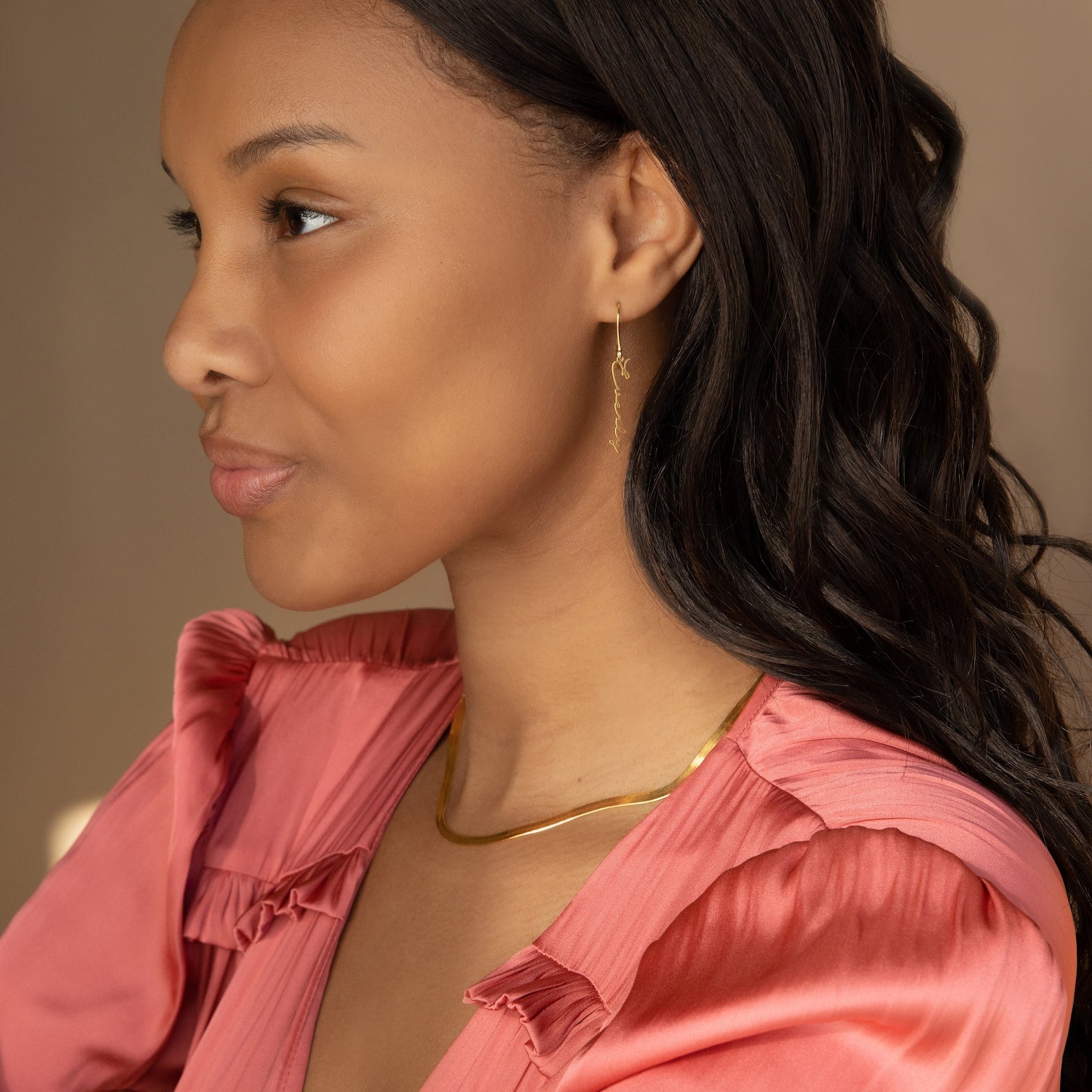 A woman in a pink blouse wearing Minimal Dangling Name Earrings and a gold necklace looks to the side—these earrings make an ideal bridesmaid gift.