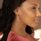 A woman in a pink top is shown in profile against a neutral background, wearing the Minimal Dangling Name Earrings.