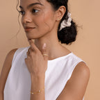 A woman wearing a white sleeveless top, cream scrunchie, and gold Grace Name Earrings gazes thoughtfully to the side.