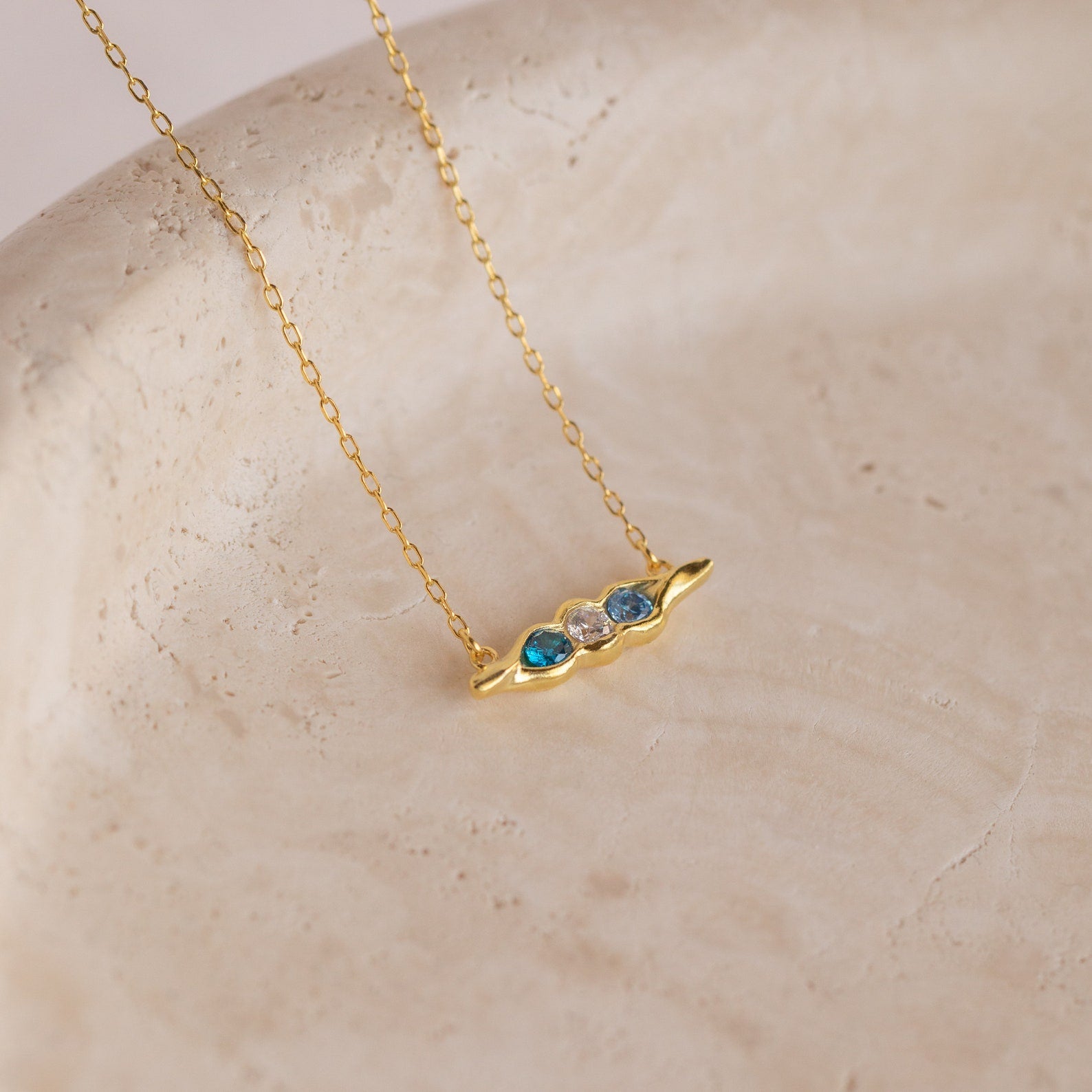 The Horizontal Pod Birthstone Necklace features a custom Peas in a Pod pendant with three blue and clear gemstones, displayed on a textured surface for a unique jewelry statement.