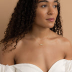 A woman with curly hair in an off-the-shoulder white top looks to the side, wearing a unique jewelry piece—the Horizontal Pod Birthstone Necklace in delicate gold.
