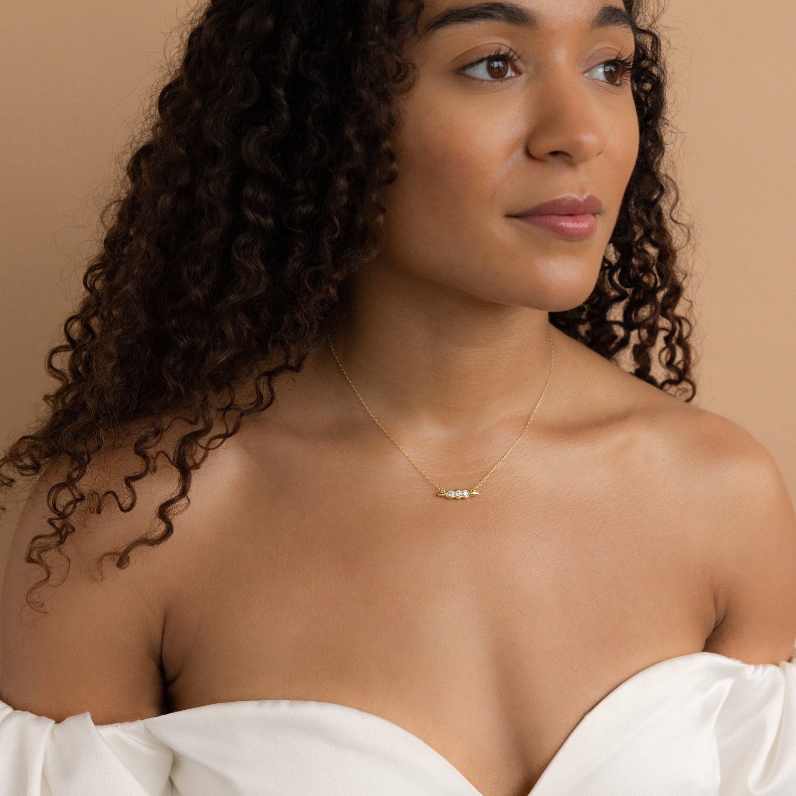 A woman with curly hair in an off-the-shoulder white top looks to the side, wearing a unique jewelry piece—the Horizontal Pod Birthstone Necklace in delicate gold.