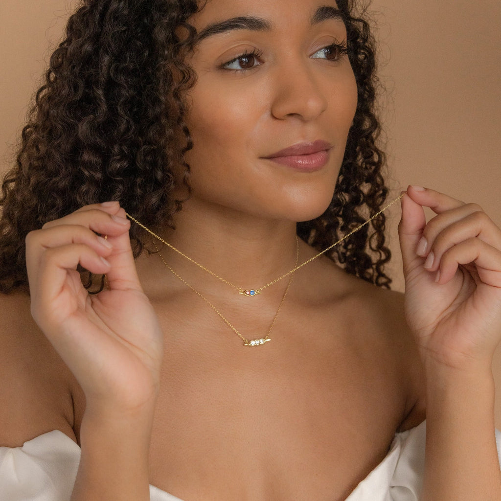 Woman with curly hair wears an off-shoulder top and layers two delicate gold necklaces, featuring the Horizontal Pod Birthstone Necklace—a unique jewelry piece with small pendants.