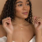 Woman with curly hair wears an off-shoulder top and layers two delicate gold necklaces, featuring the Horizontal Pod Birthstone Necklace—a unique jewelry piece with small pendants.
