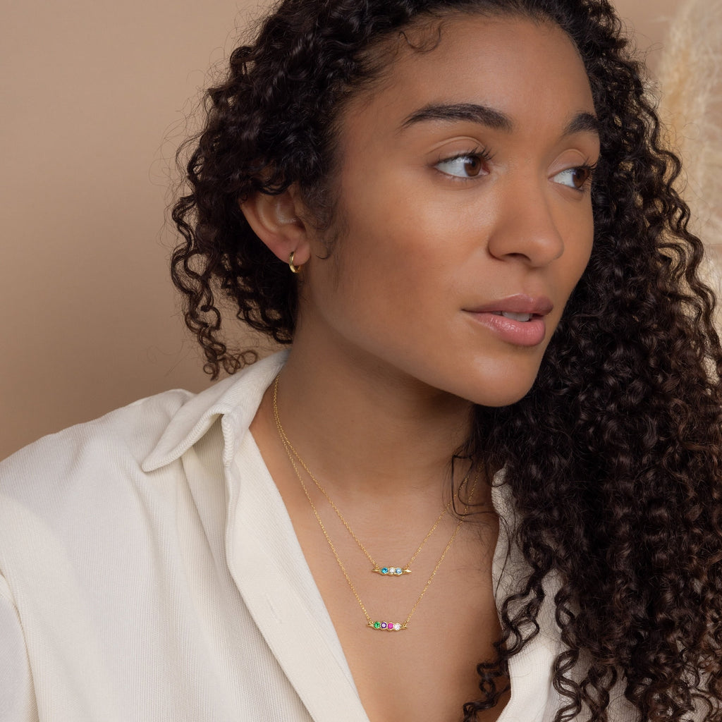 Woman with long curly hair wears a cream shirt and layered gold necklaces with colorful beads, featuring a unique jewelry piece—a Horizontal Pod Birthstone Necklace.