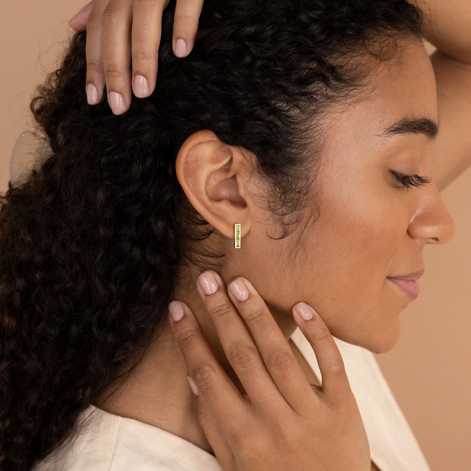 A woman with curly hair touches her earlobe, showcasing the Baguette Birthstone Bar Studs in gold against a beige background.