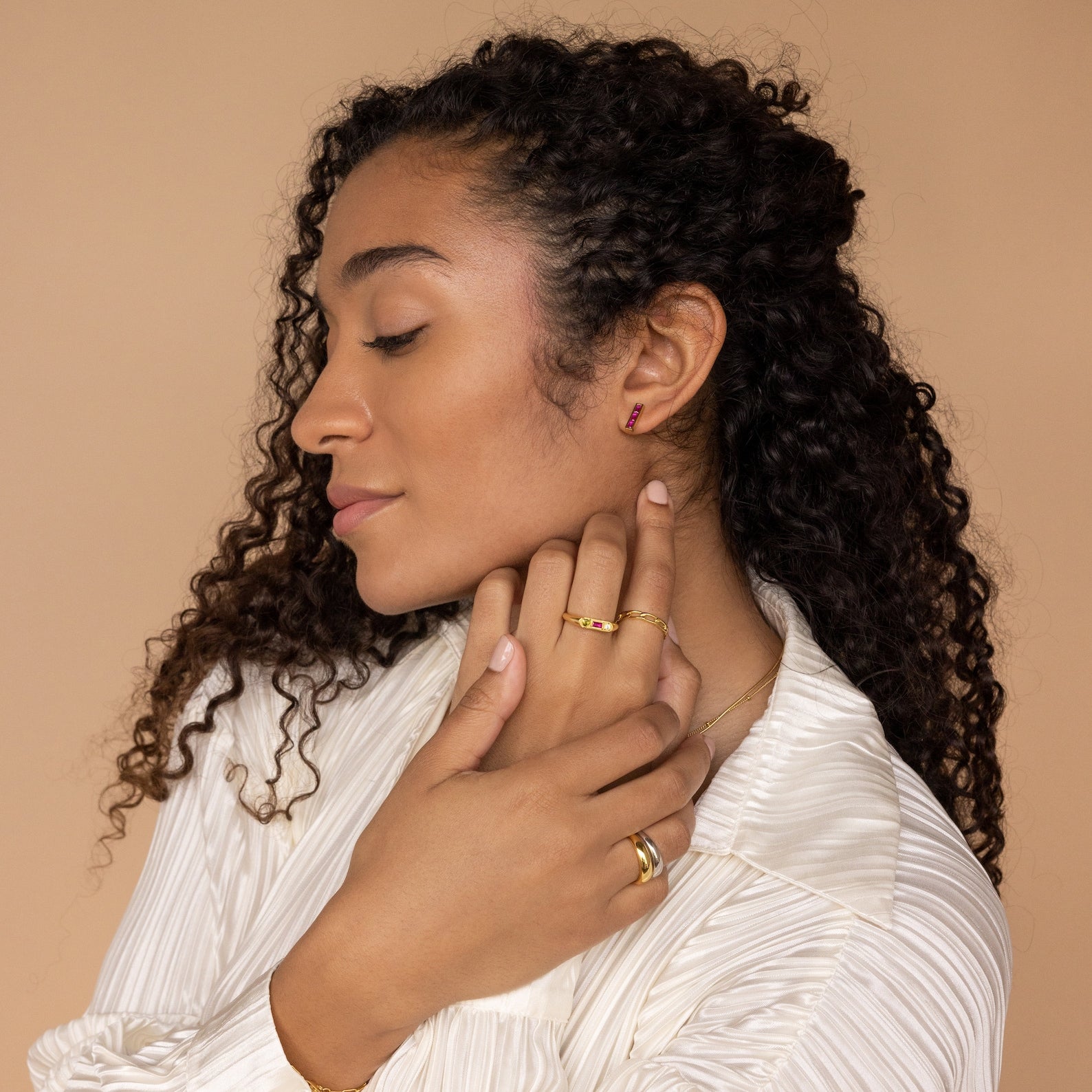 Woman with curly hair models Baguette Birthstone Bar Studs and a white blouse, eyes closed, against a beige background.