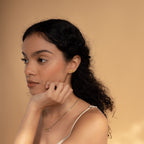 Woman with curly hair and gold jewelry poses against a beige background, resting her chin on her hand and showcasing Old English Name Earrings—ideal as a personalized custom gift.